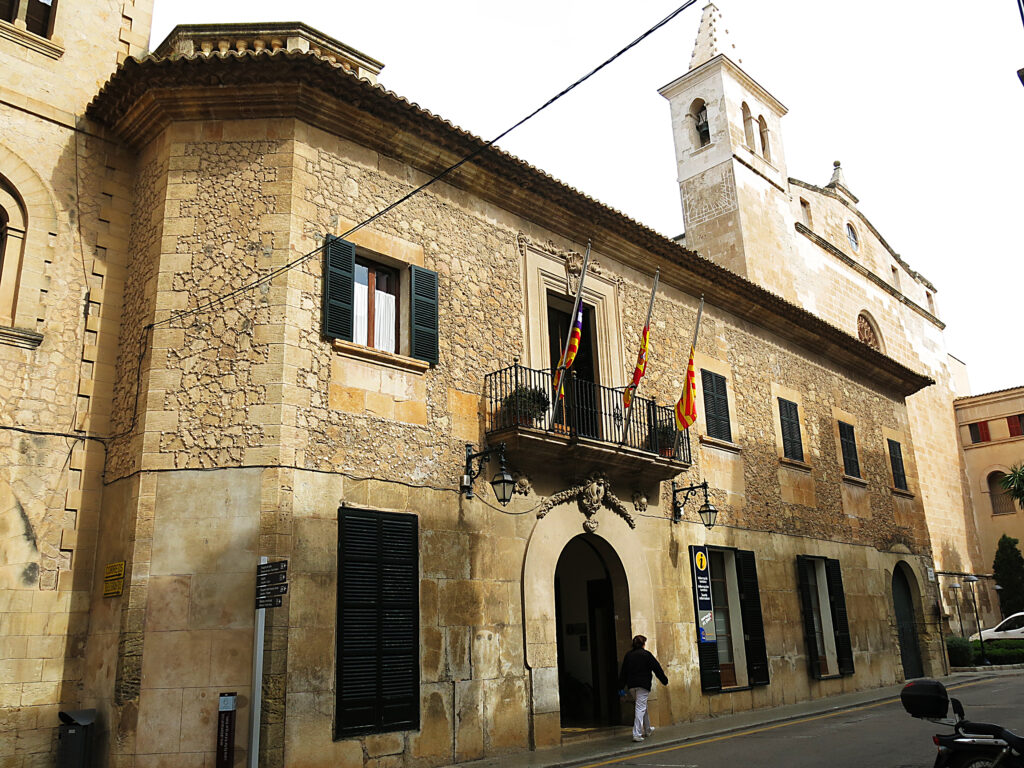 Steinfassade des Rathauses von Manacor mit Balkon und Flaggen; dahinter der Kirchturm.