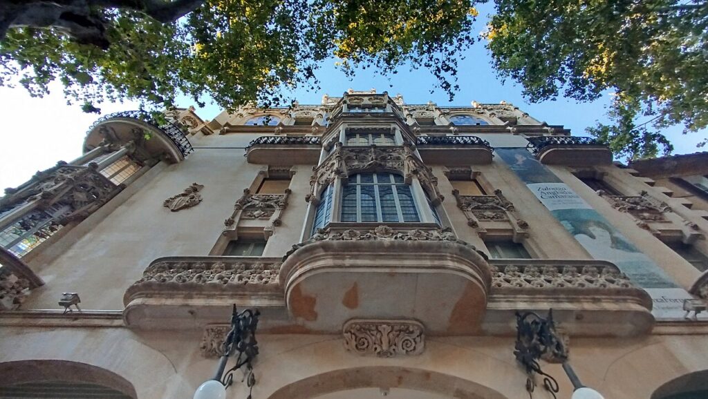 Blick von unten auf die verzierte Fassade des ehemaligen Gran Hotel in Palma, oben von Baumkronen eingerahmt.