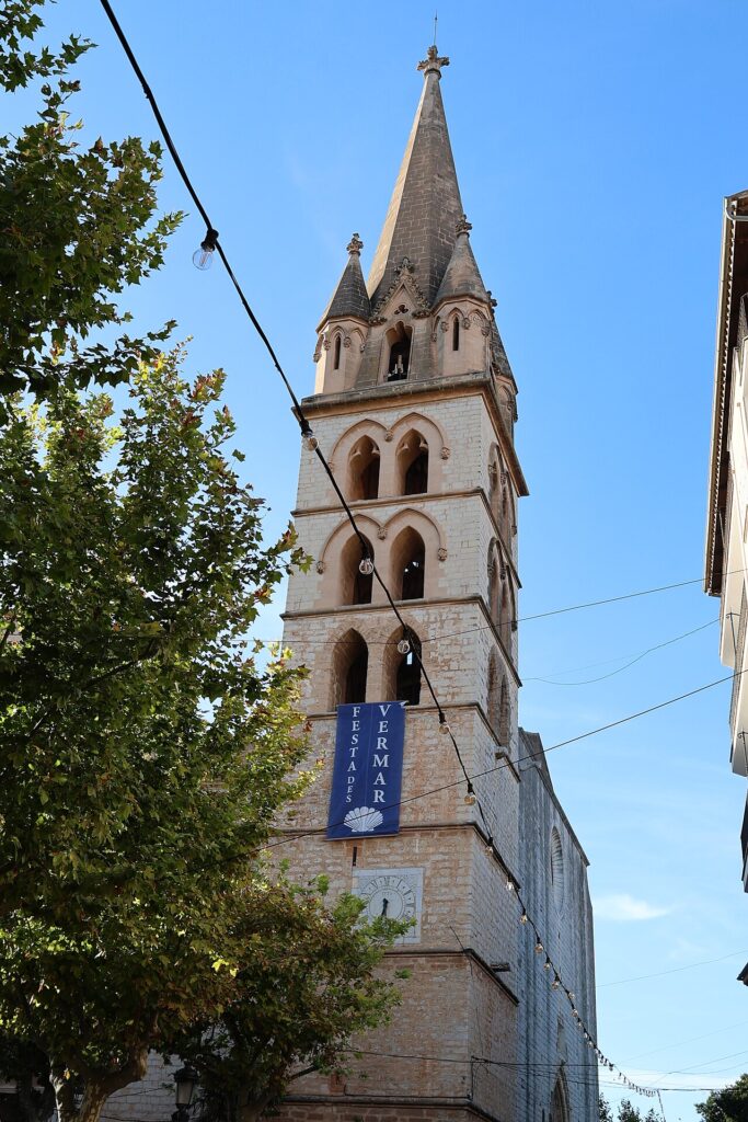 Kirchturm in Binissalem mit Vermada-Banner, Lichterketten und einem Baum im Vordergrund.