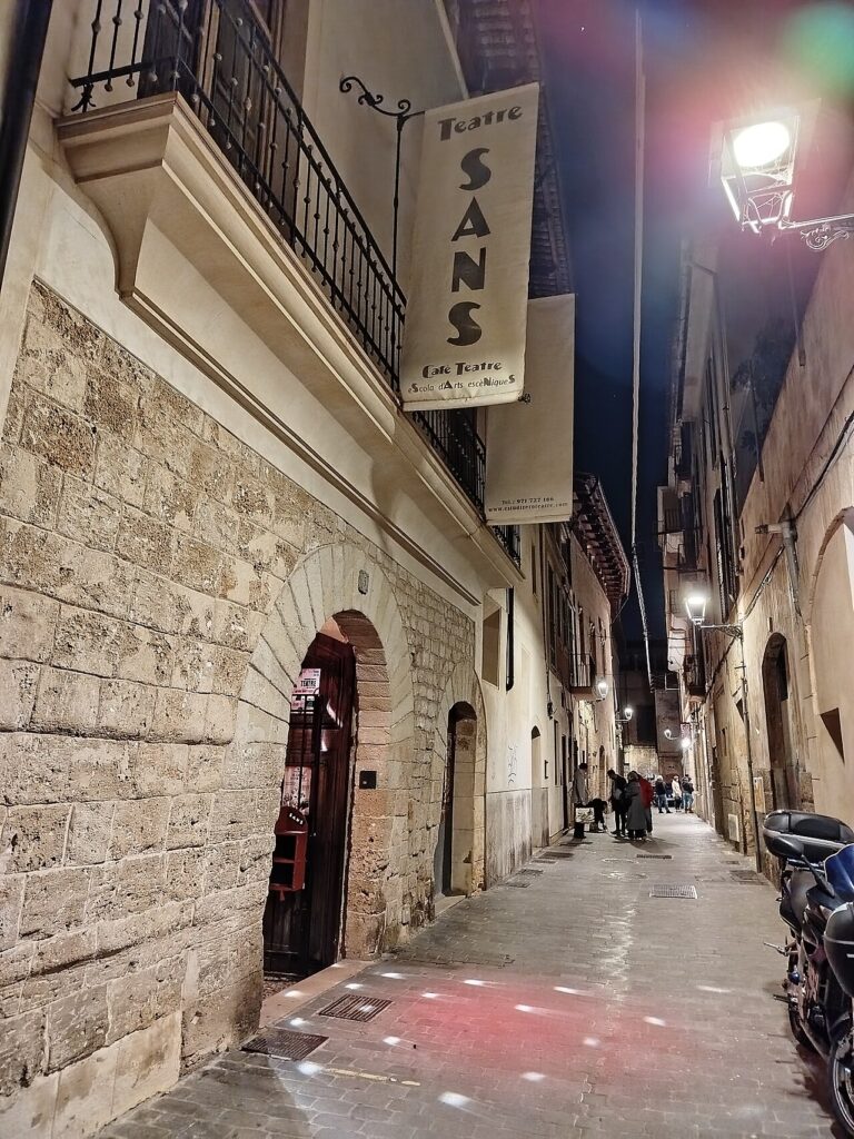 Beleuchteter Eingang des Teatre Sans mit Theaterschild in einer schmalen Altstadtgasse.